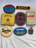 Photo of 8 vintage items including patches, pins, tins, and a bottle cap on white fabric background.