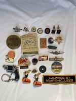 Overview of various vintage pins, badges, small toy revolver, and embroidered patch arranged on white cloth.