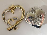 Photo showing two heart-shaped pendants, one gold-tone with clear rhinestones and a smaller heart inside, and one silver-tone with a crowned heart design.
