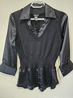 Front view of a black ladies top hanging on a hanger, showing satin collar, V-neckline, 3/4 sleeves with satin cuffs, textured fabric body, and button detail at peplum hem.