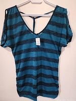 Front view of a teal and black horizontally striped ladies top with cold shoulder cutouts hanging on hanger against plain wall.