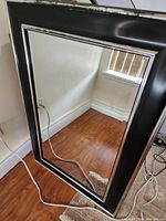 Framed wall mirror resting against a wall on a wooden floor, showing full front view of mirror and frame detail.