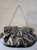 Full front view of metallic black purse showing gathered pleated design and chain strap on a beige carpet.