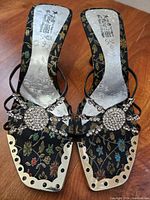 Pair of black fabric women's sandals with floral pattern, jeweled rose embellishment on straps, chromed metal front edge, size visible as 6 1/2, insole with embossed floral design.
