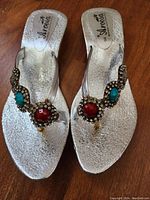 Pair of silver metallic sandals with jeweled embellishments on wooden floor.