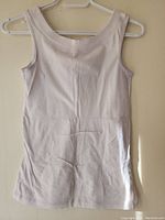 White sleeveless tank top hanging on a hanger against beige wall, back view, showing entire garment.