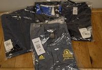 Four Adidas Boston Marathon apparel items in original sealed packaging: a blue half-zip jacket, a grey polo shirt, a blue and grey long sleeve shirt, and a black polo shirt.