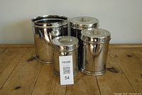 Four reflective stainless steel canisters displayed on a wooden surface, varying in size, with one missing its lid.