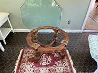Octagonal glass top removed from wooden base, placed standing against wall. Wooden base positioned on small rug with red and white floral pattern. Floor covered with dark hexagonal tiles.