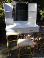 Full view of the white dressing table with gold metal legs, squared mirror with light bulbs, and shelving on both sides. Matching fabric-cushioned stool in front.
