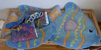 Two blue fish-shaped bath mats with orange, yellow, and purple wavy patterns, along with folded ocean/fish themed curtains and accessories.