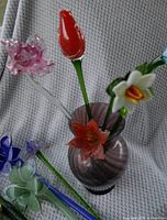 Overall view of multiple glass flowers with varied colors including red, white, pink, orange, and blue in a purple swirl glass vase.