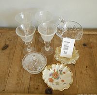 Set of 6 clear wine glasses, pedestal bowl with handle, clear covered candy dish, scalloped clear glass candy dish, floral decorative plate on wooden surface