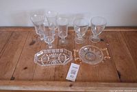 Set of 5 clear stemmed glasses, 1 rectangular cut glass plate, and 1 round serving dish with gold handles arranged on wooden table.