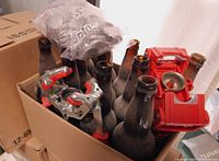 Cardboard box with used glass beer bottles, red handheld bottle capper, chrome-mounted bottle capping tool, and a plastic bag with metal bottle caps labeled 'BOCK'.