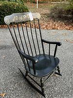 Black wooden rocking chair with vertical spindle back, floral painted top backrest, and curved arms, shown outdoors on pavement with leaves around.