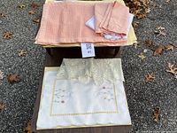 Photo of assorted table linens including peach and yellow placemats, napkins, and lace doilies placed on a small table outdoors on asphalt ground with fallen oak leaves