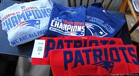 Grey NFL Super Bowl Champions hoodie, navy Super Bowl Champions sweatshirt, navy short sleeve T-shirt, and two navy/red Patriots scarves on table
