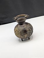 Bronze vase with raised dragon and floral decoration, visible handle with animal head mounts, and three legs, photographed on black background to highlight details.