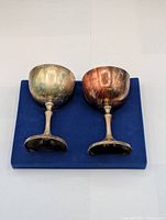 Pair of small silver-tone goblets shown side by side on a blue background, highlighting the tarnished and aged metal surface.