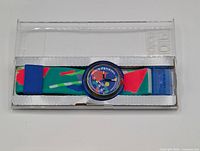 Swatch wristwatch with colorful fabric strap and blue bezel inside clear plastic case, front view.
