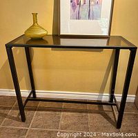 Front angle view of the glass side table showing the cappuccino brown metal frame and clear glass top with a yellow decorative vase and framed print behind it.