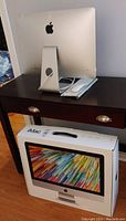 Apple iMac on table with original box underneath. Shows iMac from back with keyboard and mouse placed on table.