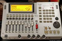 Top front view of Korg D12 digital multitrack recorder showing 12 channel faders, buttons, numeric keypad, jog dial, LCD screen, and label indicating version 2.00 software. Device is silver-grey with black and red controls.