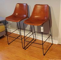 Two light tan molded leather high chairs with black metal base shown side by side against a wall on hardwood floor.