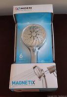 Front view of boxed Moen Magnetix shower head showing the chrome shower head and packaging with blue background.