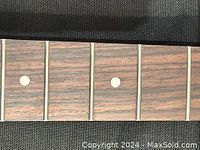 Close-up image of the rosewood guitar neck fingerboard showing dot inlays and metal frets on a dark wood grain surface.