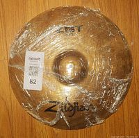 Photo of a pair of Zildjian ZBT high hat cymbals wrapped in plastic on a wooden floor, showing logos and general condition.