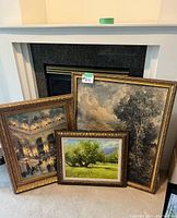 Three framed artworks leaning against a fireplace: one painting, two prints all with ornate gold frames.