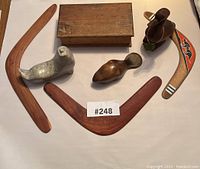 Overview of the 3 wooden boomerangs and 3 animal carvings including metal seal and wooden bird with box in background.