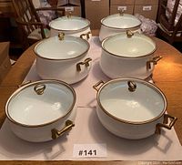 Overhead view of six white round cookware pieces with brass handles and glass lids arranged on table.
