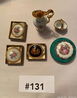 Six pieces including Limoges brooches in ornate gold frames, small decorative jug, heart-shaped ring holder, and a round trinket box arranged on a beige surface