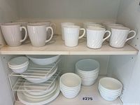 Wide view of a kitchen cupboard shelf containing 10 white mugs on the top shelf, various plates, and bowls on the lower shelf including multiple stacked bowls and plates.