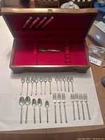 Wooden silverware box open showing red velvet interior, 4 stainless knives in lid rack, butter spreader in base, sterling flatware laid out on paper