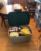 Large Rubbermaid bin on dining table filled with folded clothing including plaid shirts and yellow knit garments.