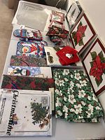 Multiple Christmas tablecloths, placemats, napkins, and oven mitts arranged on a table with framed poinsettia prints in the background.