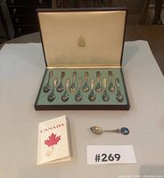 Box with 12 Canadian Province souvenir spoons and one additional spoon with blue enamel emblem, plus informational booklet and lot number card.