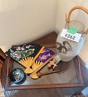 Photo showing all lot items on a wooden table corner: pottery beverage dispenser with spigot, two folding fans, small round black lacquer box, wooden and glass serving tray frame.