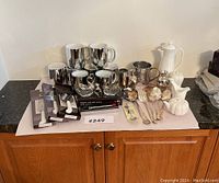 Overview of the entire lot showing silver plated mugs, serving spoons and forks, ceramic pitchers and coffee pot, silver plated boxed knives, and other assorted silver plated items.