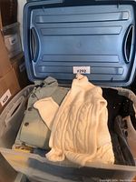 Top view of large blue plastic bin with light jackets and sweaters folded inside, showing cream cable knit sweater and green jacket with buttons