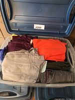 Plastic bin with several folded clothing items including pants and jackets in various colors such as beige, orange, olive green, and maroon.