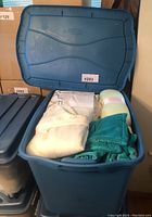 Open blue Rubbermaid bin containing folded white garments, teal velour zip-up jacket with tag, and pastel green and white bolster pillow tied with a satin ribbon.
