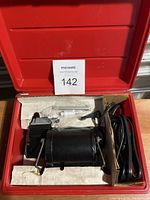 Black 12V car air pump stored in red carrying case without hose, showing power cables and foam padding.