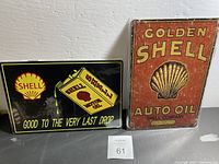 Photo of two new decorative tin signs featuring Shell Oil branding. One sign shows a black background with a tilted oil can pouring oil and the slogan 'Good to the Very Last Drop'. The other sign has a rusty orange background with a shell logo and the text 'Golden Shell Auto Oil Shell Company'.