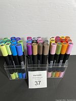 Top-down view of three organized clear plastic holders containing 34 dual tip artist markers with colorful caps arranged by color.
