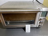Front view of Breville Convection Smart Oven showing stainless steel finish, glass door, and handle. A MaxSold lot number card is visible in front of the oven.